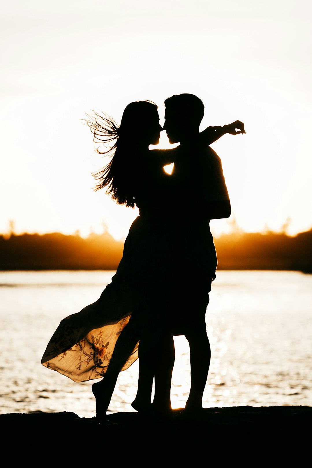 Casal em silhueta, nariz com nariz, em contraluz ao pôr-do-sol junto à água